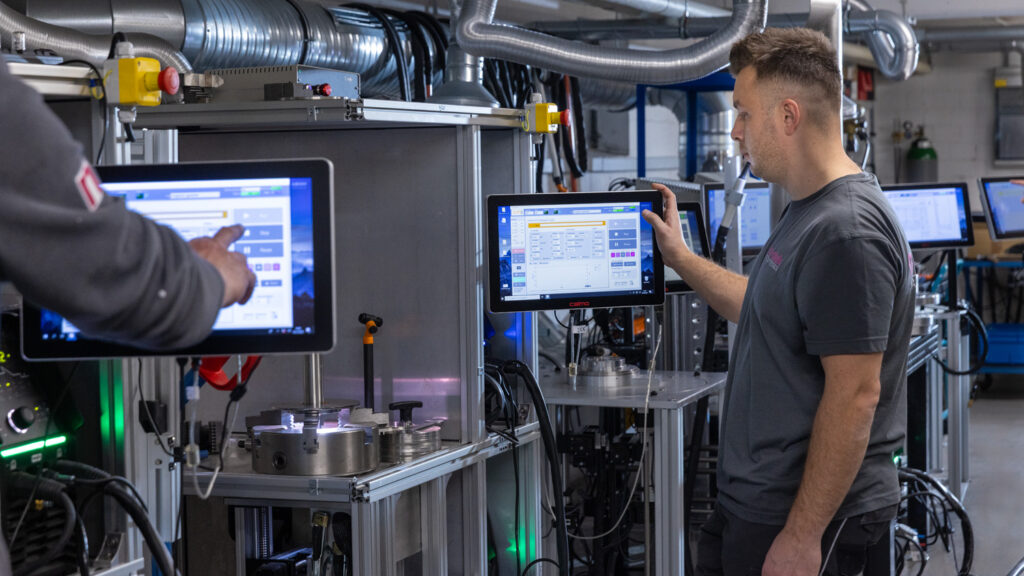 Mitarbeiter stehen an modernsten Schweißrobotern und geben Daten via Tablet ein